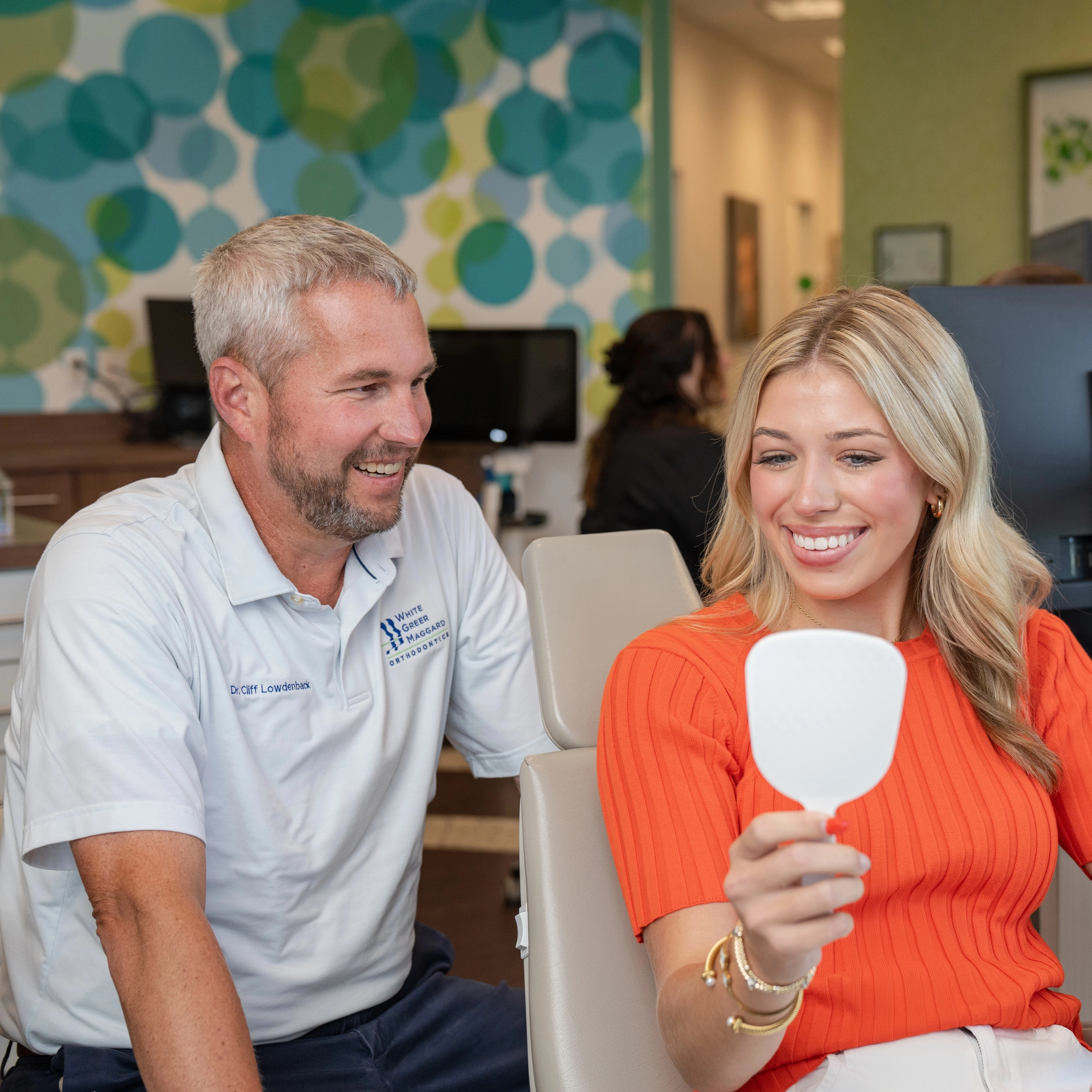 A happy, blonde woman with a perfect smile looks at her reflection in a small white handheld mirror. She is wearing an orange long-sleeved shirt and gold hoop earrings. Behind her, a dental office is visible, including another person and a computer monitor.